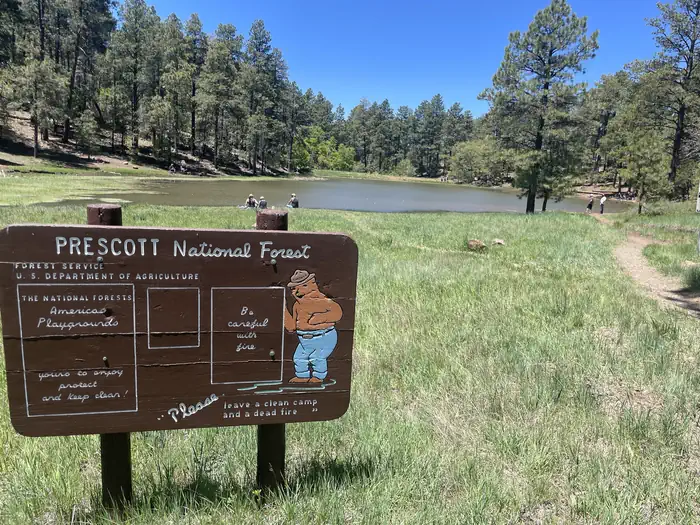 A photo of Mingus Lake including a walking path, people on the shoreline and a Prescott National Forest sign featuring Smokey Bear.