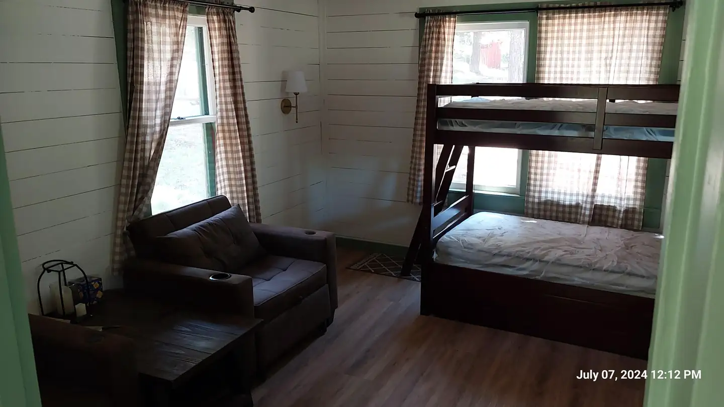 Bunk beds and chairs in the main living room seen from the kitchen. The scene is dimly lit as sunlight enters the room through 3 large windows.