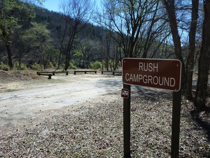 A brown sign stating "Rush Campground" can be seen in the foreground with a dirt road, trees, and a parking area in the background.