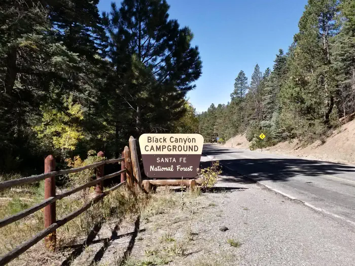 A campground sign with the words Black Canyon with a road and trees in the background.