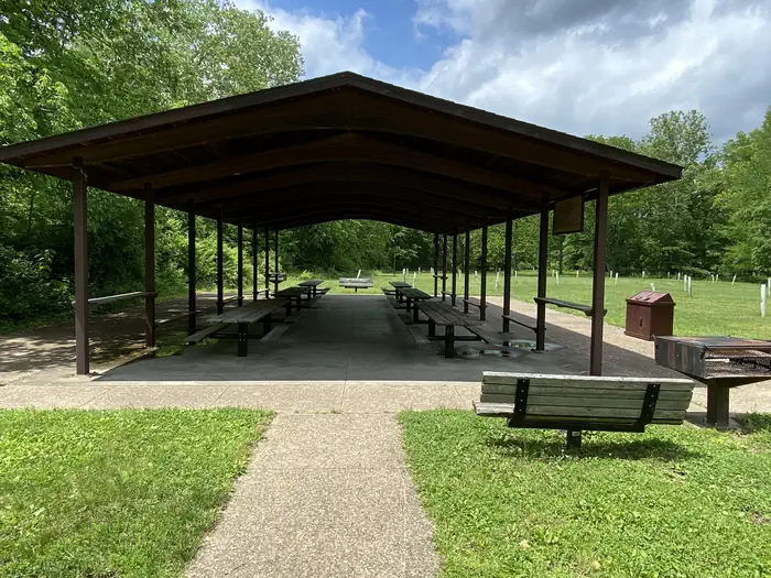 A view of an open sided pavilion with picnic tables, pedestal BBQ grill, and benches with a background of green vegetation.