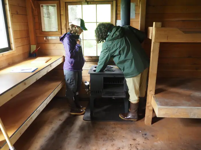 Two people lighting the stove in Gut Island Cabin 2 with wooden bunkbeds and cabinets