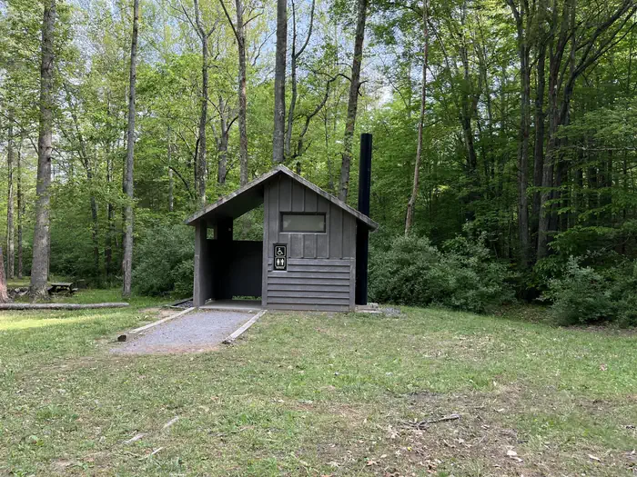 A gray vault toilet facility with a gravel ramp