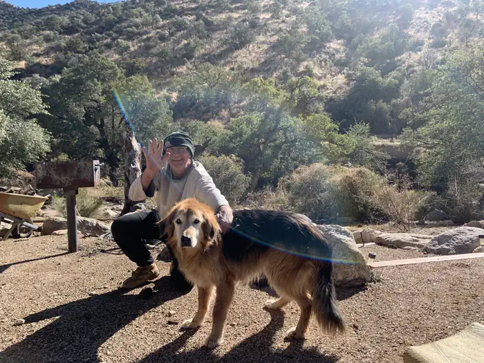 A man squatting down next to his dog, the man is smiling and waving at the camera.