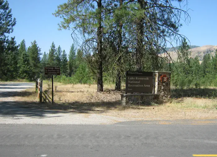 The photo shows a stone‑based National Park Service entrance sign for Lake Roosevelt National Recreation Area (Evans) standing among tall pines.