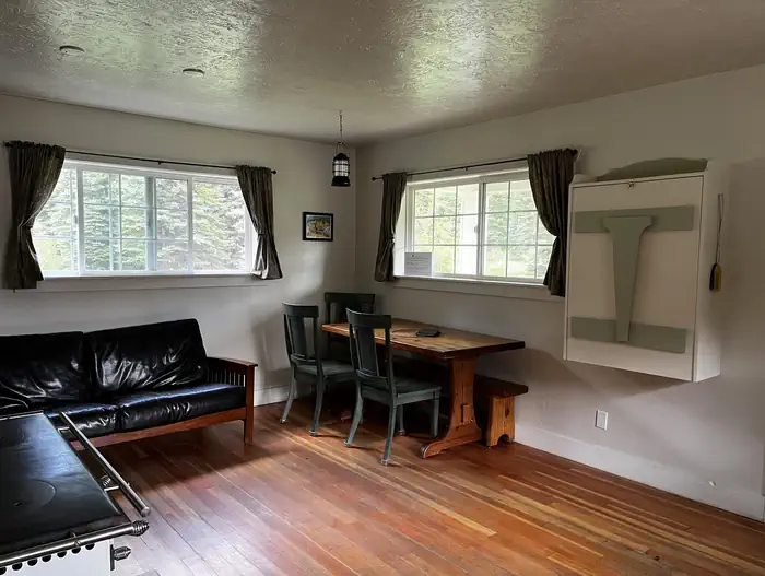 Front room of cabin showing  table, chairs, couch and two windows.