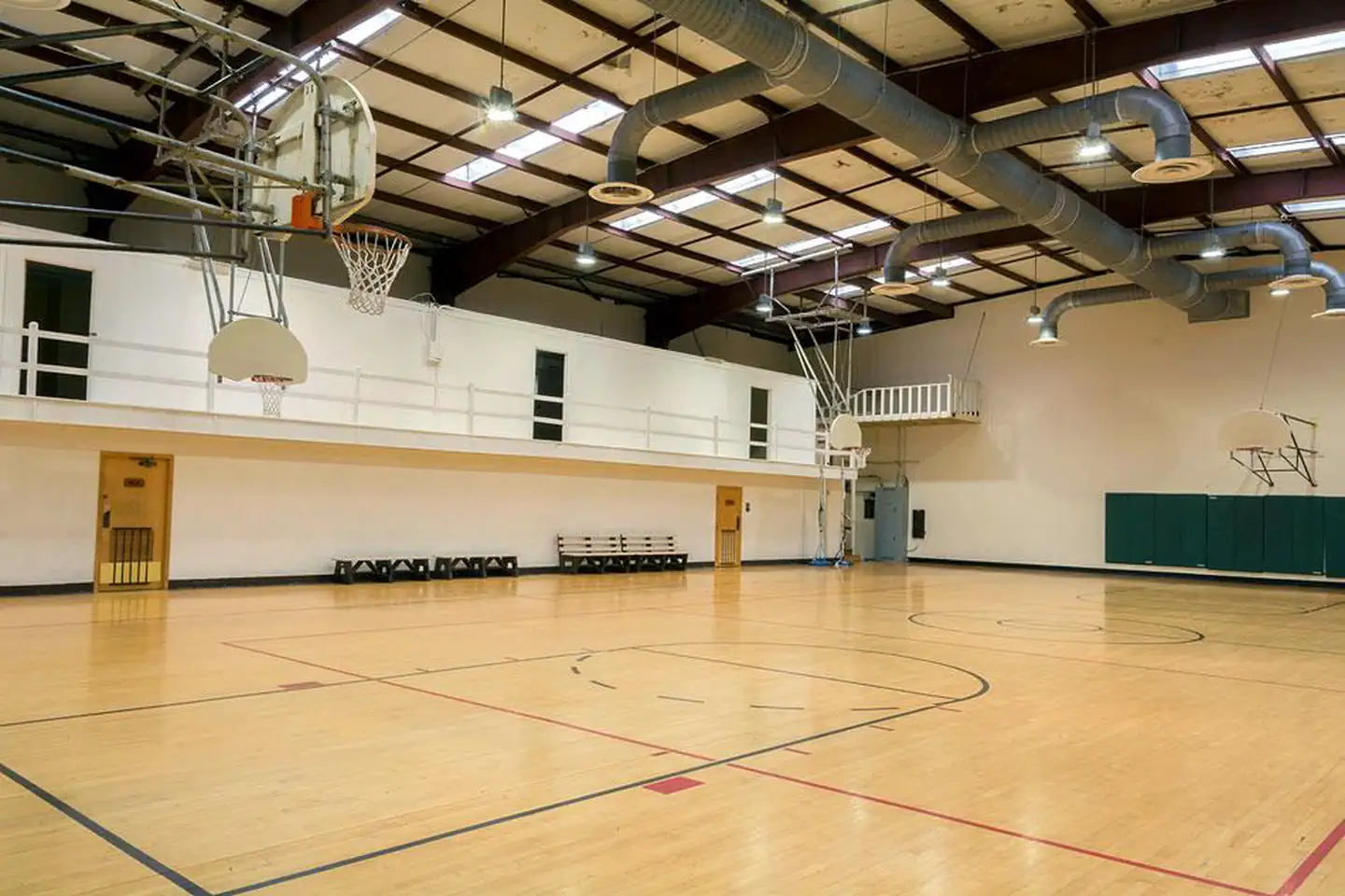 Interior of gym.  Wooden marked basketball court with 4 backboards/nets and 4 benches on sidelines. Metal beam ceiling with skylights. 3 doors visible to upper classrooms.