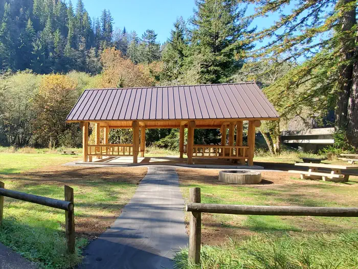 Large flat field.  Tents and picnic tables in front of a road and large conifer treein the back, fire ring and picnic table behind single pole fence in foreground.